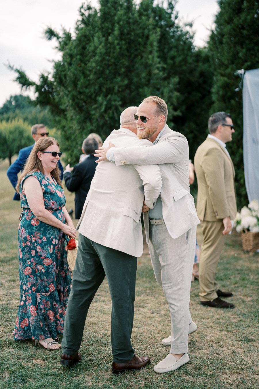 Guests in formal attire exchanging hugs and greetings outdoors at a wedding ceremony, with greenery in the background.