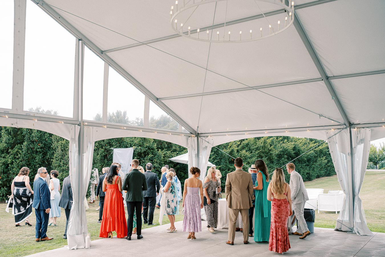 Outdoor event with elegantly dressed guests under a white tent, surrounded by greenery.