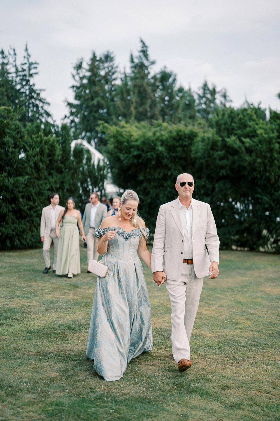 Elegant wedding guests in pastel attire walking on a lawn, with lush greenery and a group in the background.