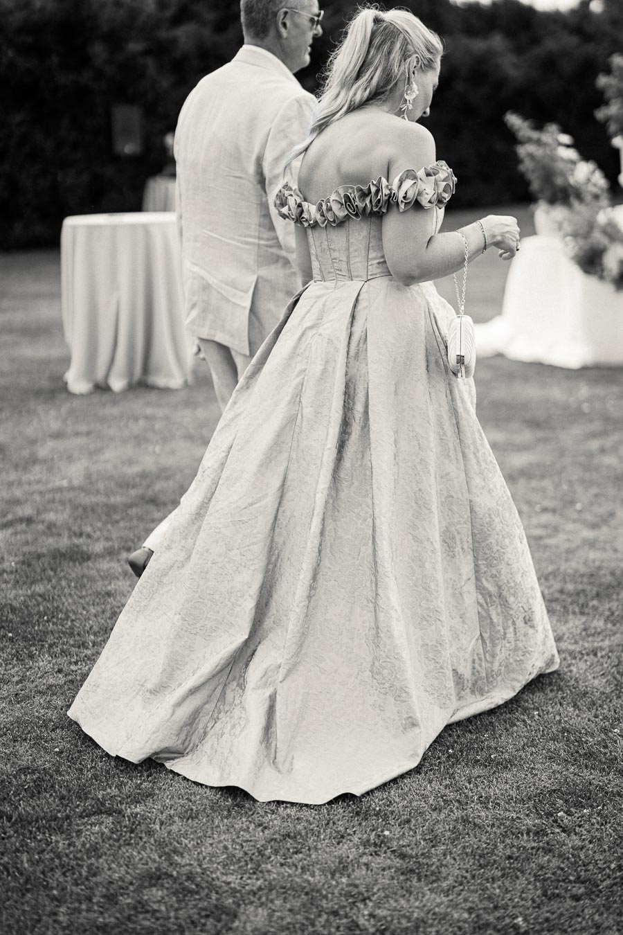 A woman in an elegant, off-the-shoulder gown, walking alongside a man in a light suit at an outdoor event. The scene captures a sophisticated and timeless moment in a black and white photo.