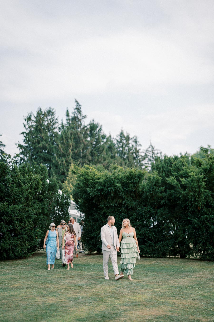 A group of elegantly dressed people walking on a grassy field surrounded by lush green trees, with a clear blue sky overhead, suggesting a daytime outdoor event.