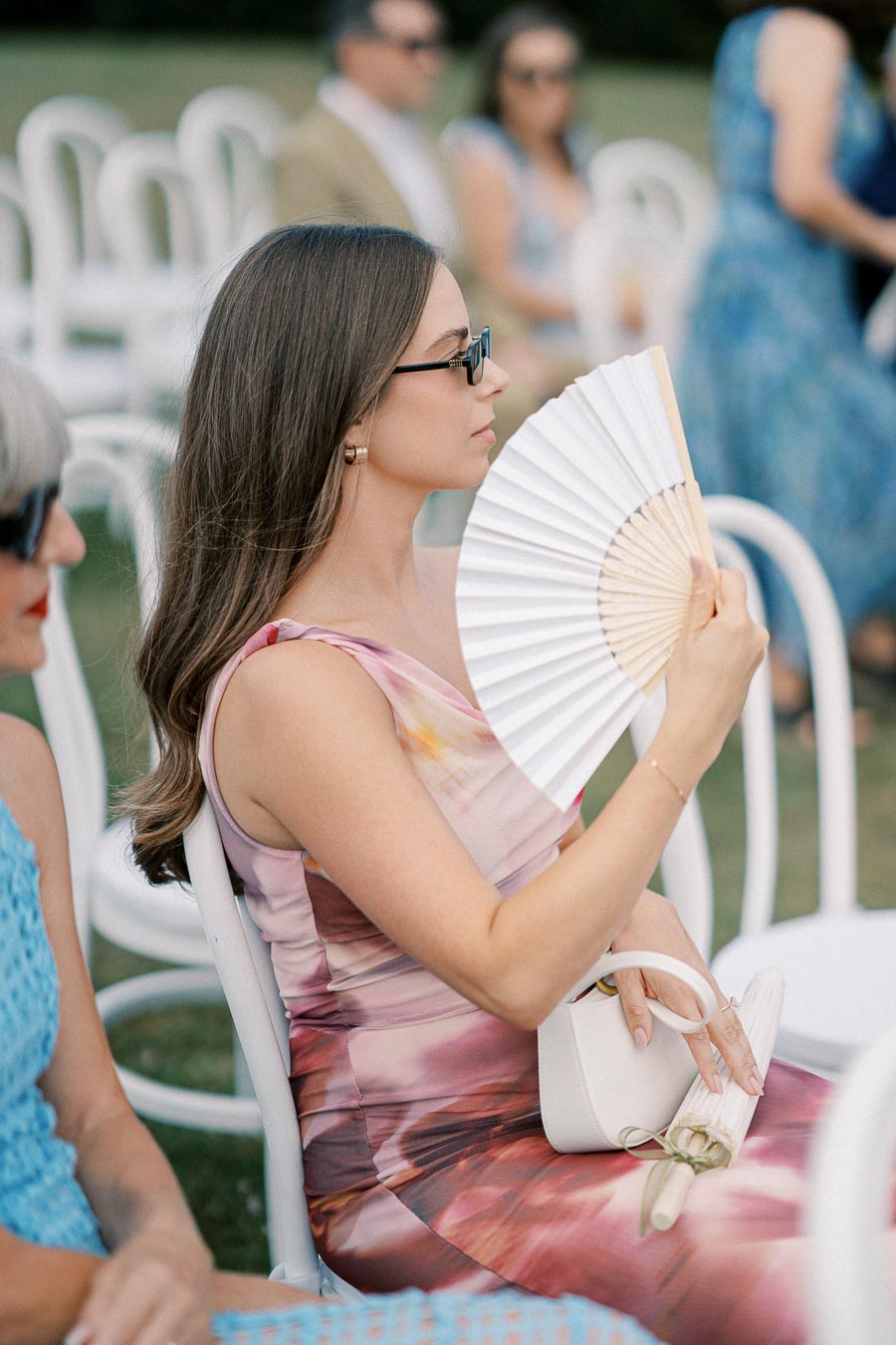 A woman in a colorful dress uses a handheld fan while seated at an outdoor event.