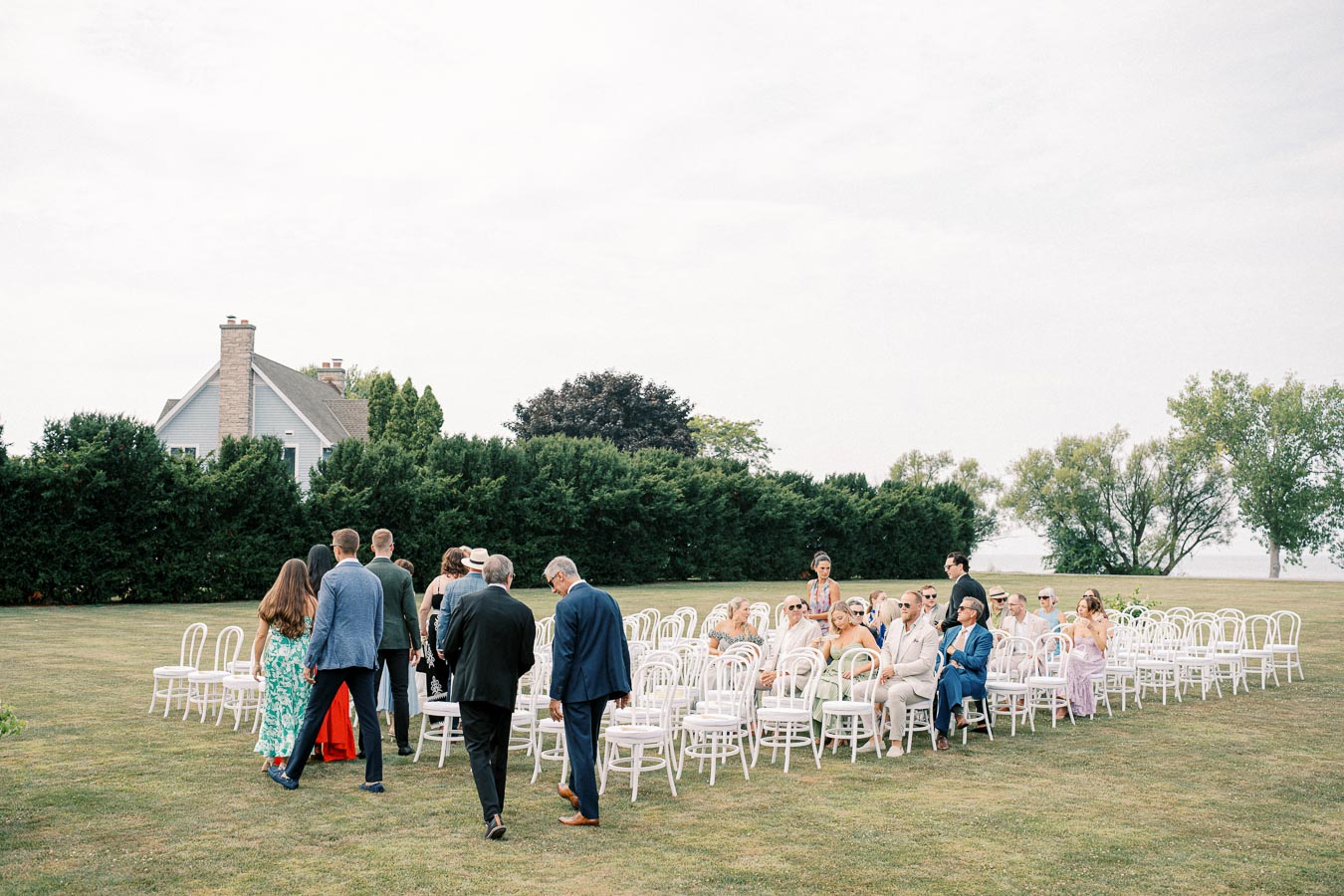 An outdoor wedding ceremony with guests seated on white chairs in a garden setting, surrounded by lush greenery and an elegant house in the background, under a clear sky.