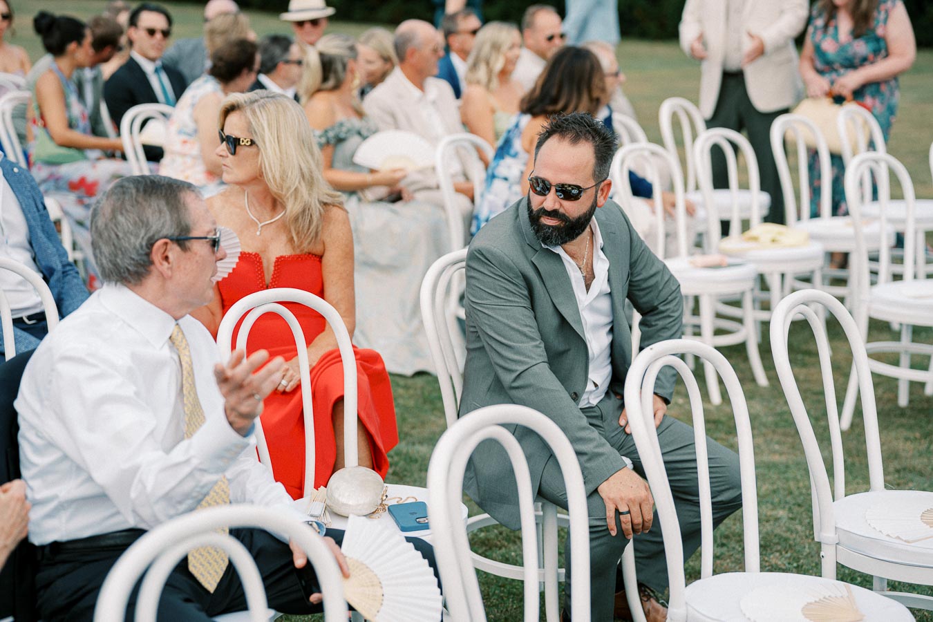 Outdoor wedding guests sitting in white chairs, dressed in formal attire, with one person in a red dress and another in a gray suit conversing, surrounded by others in elegant summer outfits.
