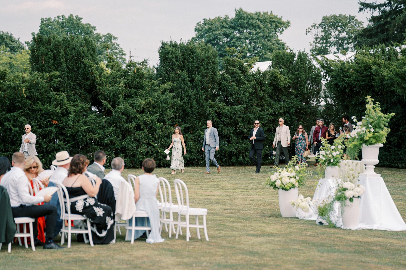 Outdoor wedding ceremony with guests seated on white chairs, lush greenery background, and attendees arriving in formal attire, enhancing the elegant garden setting.