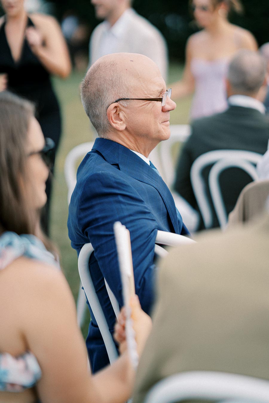 Senior man in a blue suit sitting at an outdoor event with people mingling in the background.