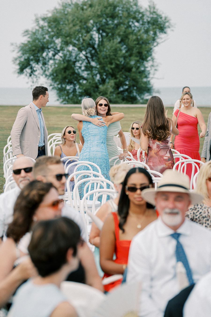 Outdoor wedding ceremony with guests in stylish attire, including bright dresses and sunglasses. A group is gathered on white chairs, with people mingling and embracing against a scenic backdrop of trees near a waterfront.