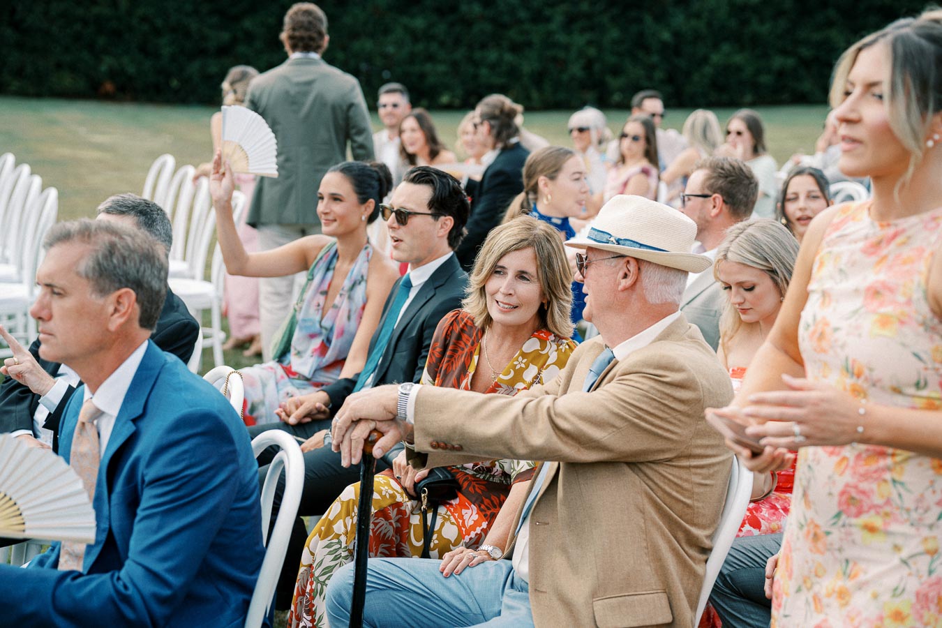 A diverse group of well-dressed guests seated outdoors at a wedding ceremony, with attendees fanning themselves and engaging in conversation amidst a sunny garden setting.