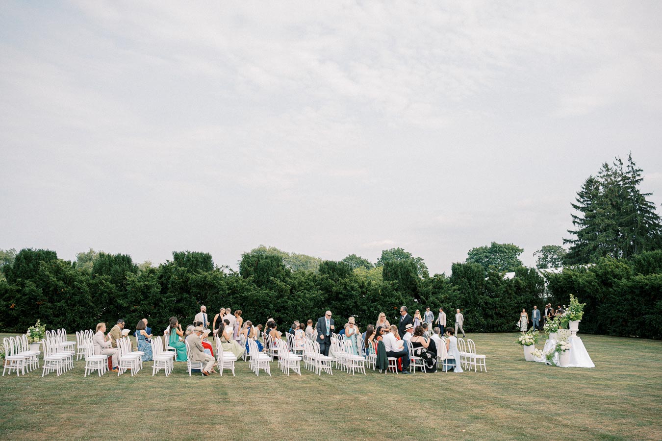 Outdoor wedding ceremony with guests seated on white chairs on a grassy lawn, surrounded by lush greenery and trees, under a cloudy sky.