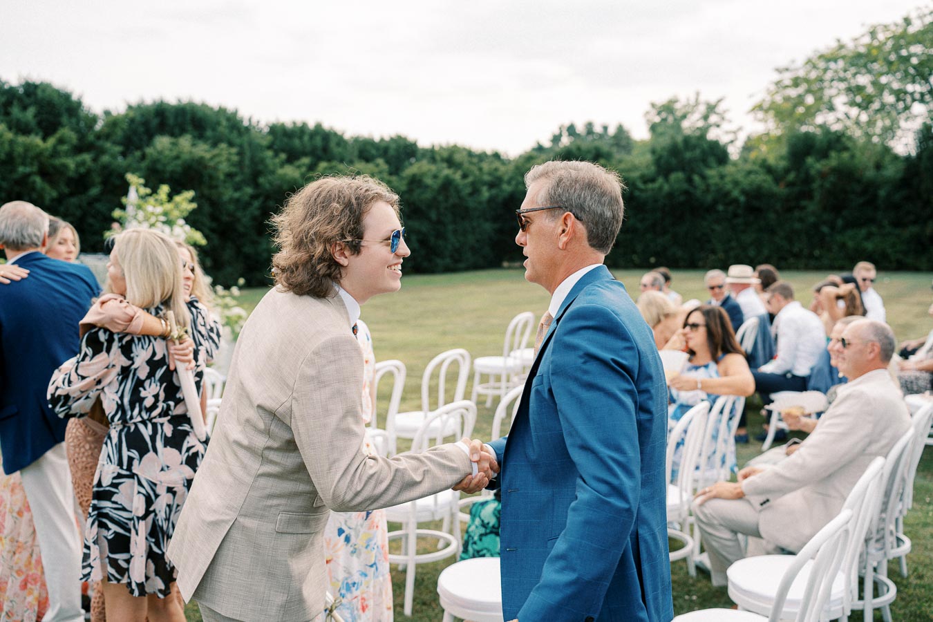 Guests greeting each other with a handshake at an outdoor wedding ceremony, surrounded by white chairs and greenery.