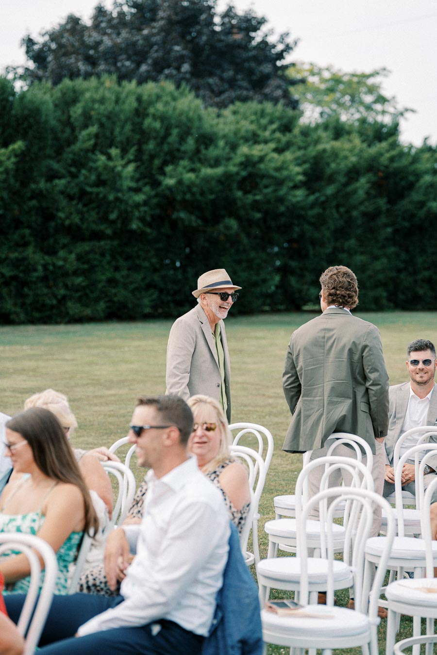 Outdoor wedding setting with guests seated on white chairs, featuring a man in a beige suit and hat standing among them on a sunny day, with green trees in the background.
