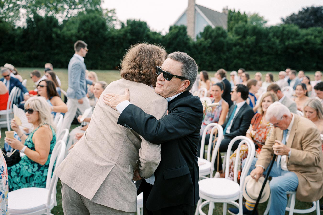 Outdoor wedding scene with two men embracing, surrounded by seated guests. The event has a formal yet relaxed atmosphere, with people dressed in suits and summer dresses, holding fans and hats. Lush greenery and a house in the background enhance the elegant setting.