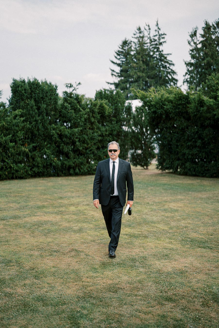 Man in a black suit and sunglasses walking on a grassy lawn with trees in the background.