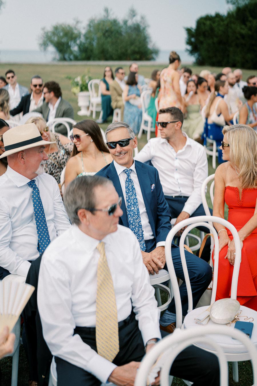 A group of elegantly dressed guests seated outdoors, some wearing sunglasses, attending a formal event with greenery and a body of water in the background.
