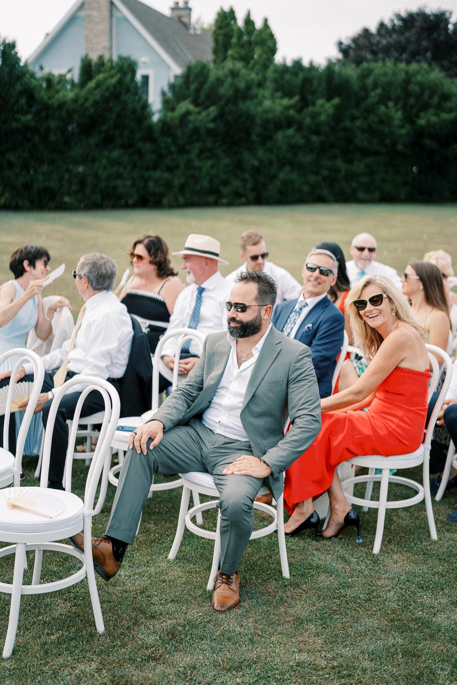 A group of elegantly dressed wedding guests seated outdoors, enjoying a sunny day with green foliage in the background.