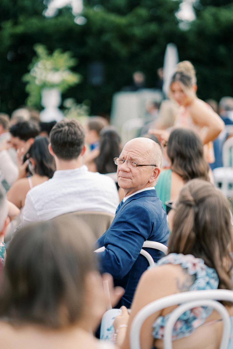Elderly man in a blue suit seated at an outdoor event surrounded by guests.