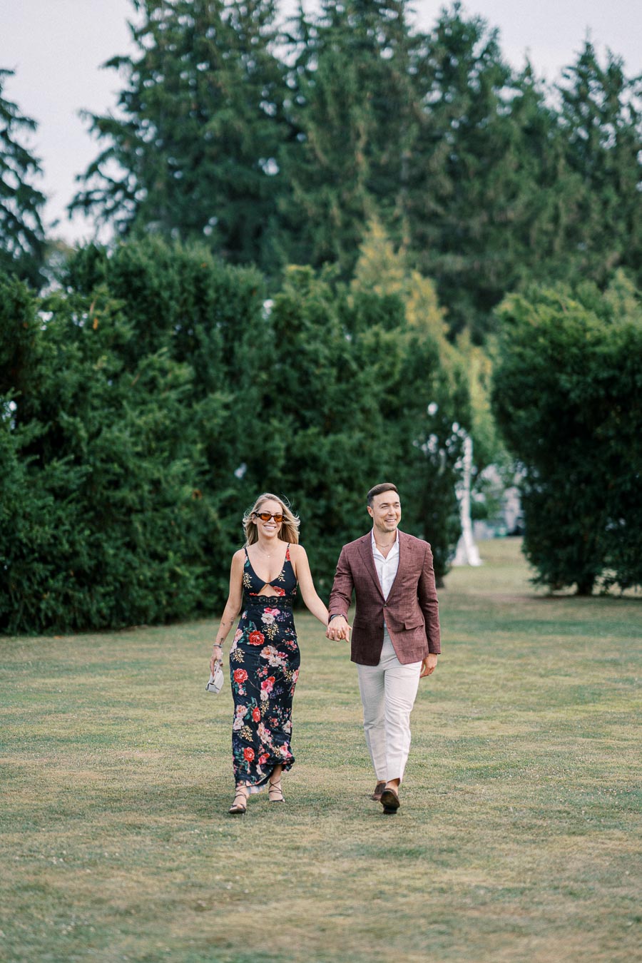A couple holding hands and walking across a grassy field, surrounded by lush green trees, with the woman wearing a floral dress and the man in a maroon blazer.