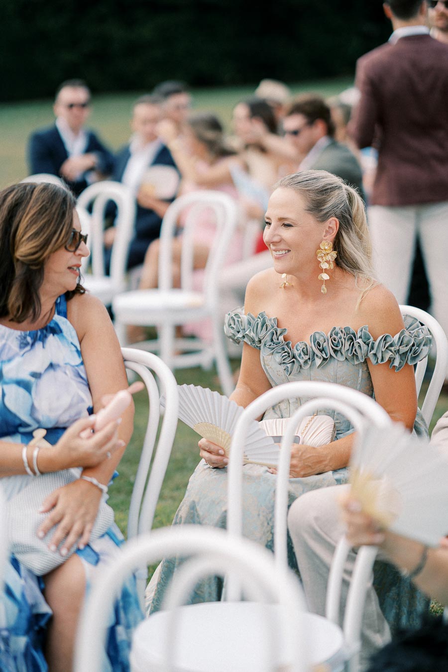 Guests enjoying an outdoor event, women seated on white chairs, wearing elegant dresses and holding fans, with others socializing in the background.
