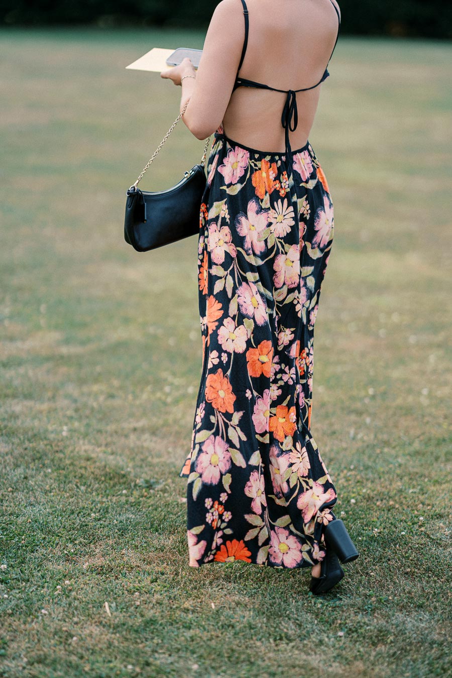 A woman wearing a stylish backless floral dress and black high-heeled shoes walking on grass, holding a black handbag.