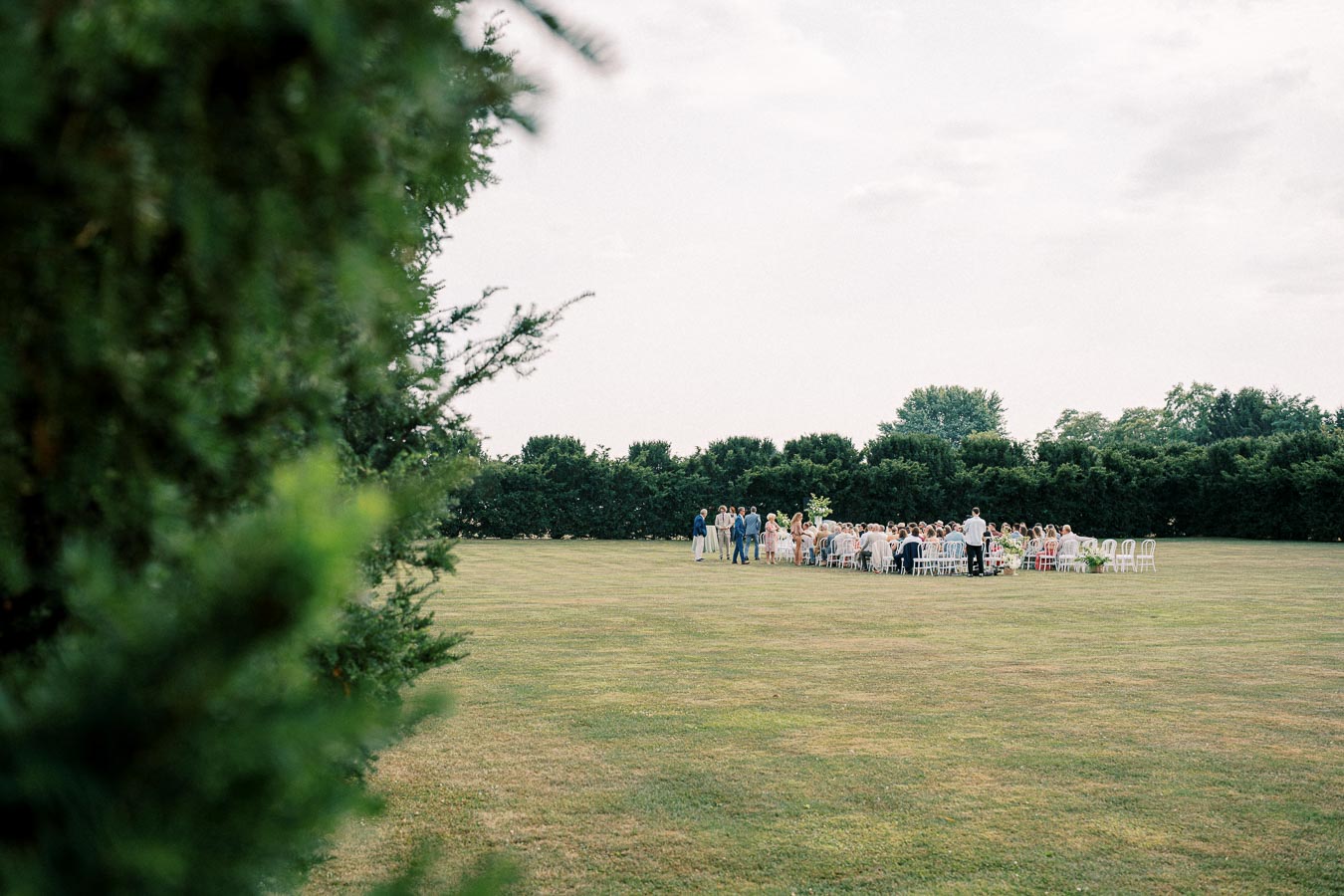 Outdoor wedding ceremony on a large grassy lawn, surrounded by lush green trees, with guests seated in white chairs under a cloudy sky.