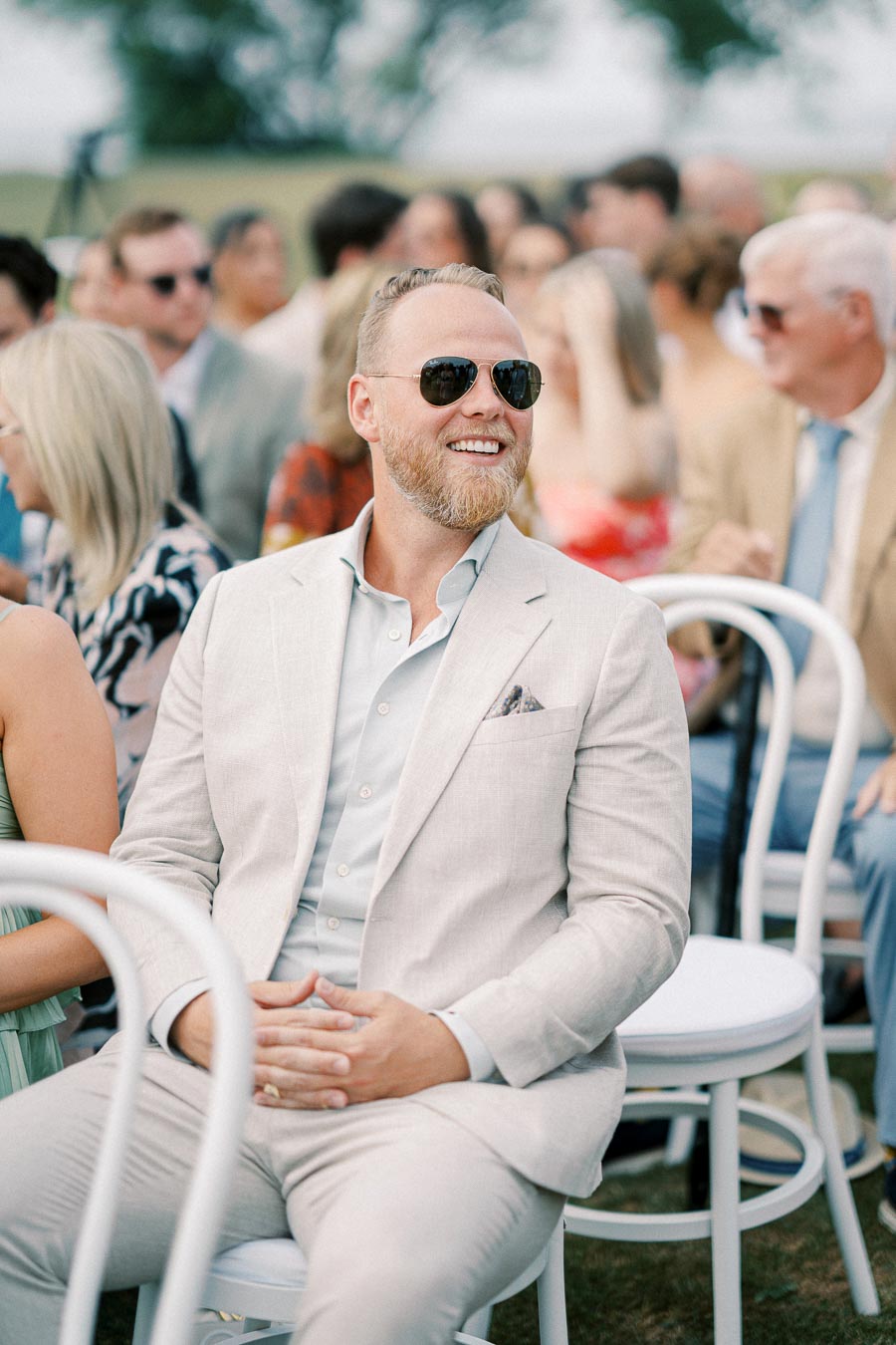 A man in a light-colored suit and sunglasses smiling while sitting on a white chair outdoors, with a blurred crowd in the background at a formal event.