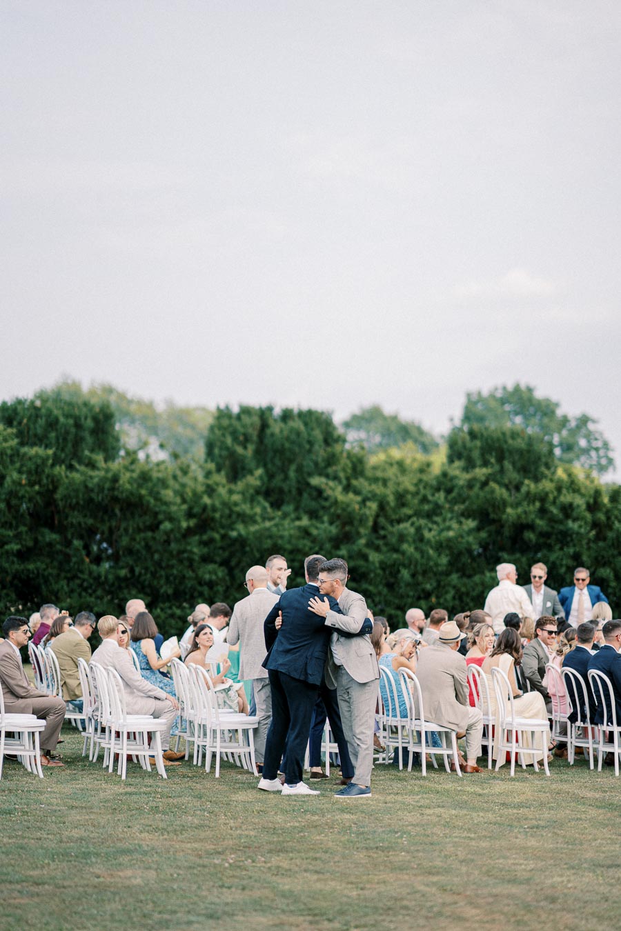 A group of well-dressed people seated and standing outdoors at a wedding ceremony, with two men embracing in the foreground and lush green trees in the background.