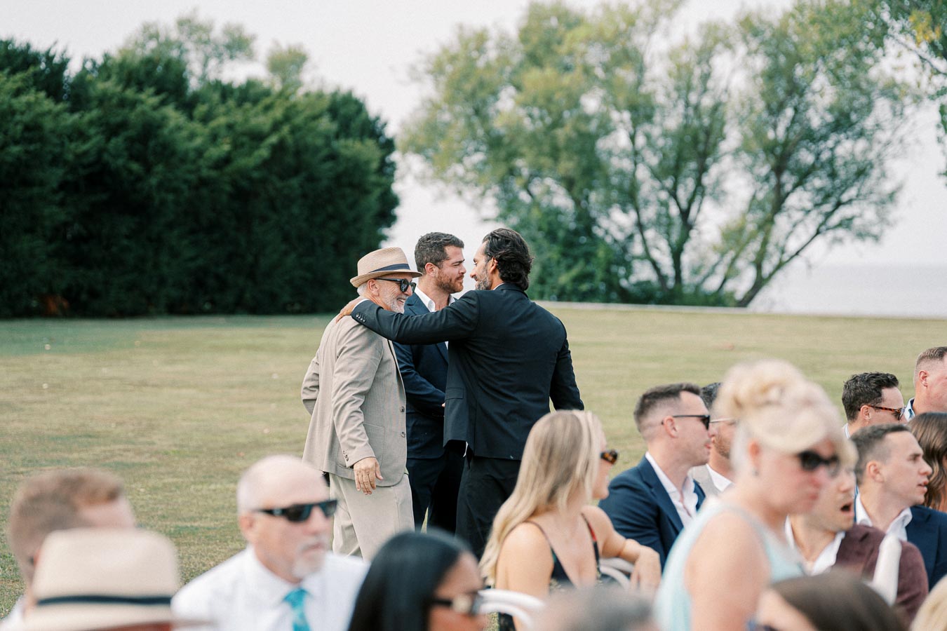 A group of well-dressed men and women socializing outdoors at an event, with lush greenery and trees in the background, creating an elegant and lively atmosphere.