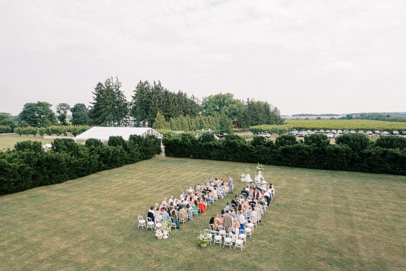 Outdoor wedding ceremony with seated guests on a green lawn surrounded by trees, a white tent in the background, and scenic views of fields and nature.
