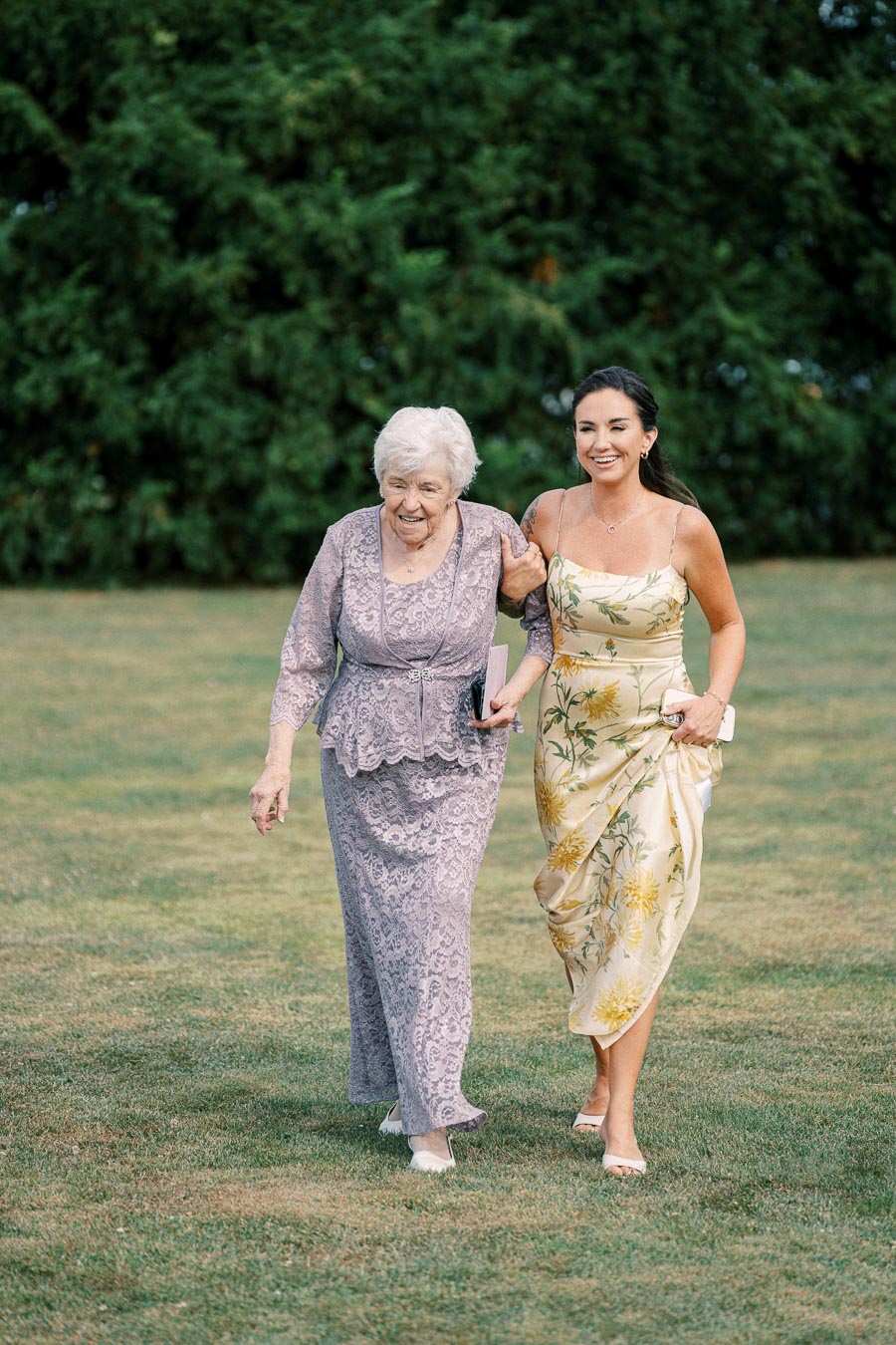 Two elegantly dressed women smiling while walking arm-in-arm on a lush green lawn, one in a patterned purple outfit and the other in a floral yellow dress.