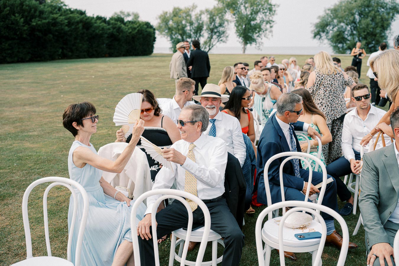 Outdoor wedding ceremony with guests seated on white chairs, enjoying a sunny day; some are using handheld fans to stay cool, dressed in formal attire, with a lush green lawn and trees in the background.