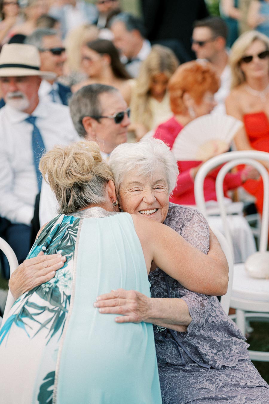 Two elderly women share a joyful embrace at an outdoor event, surrounded by seated guests in formal attire, capturing a moment of connection and happiness.