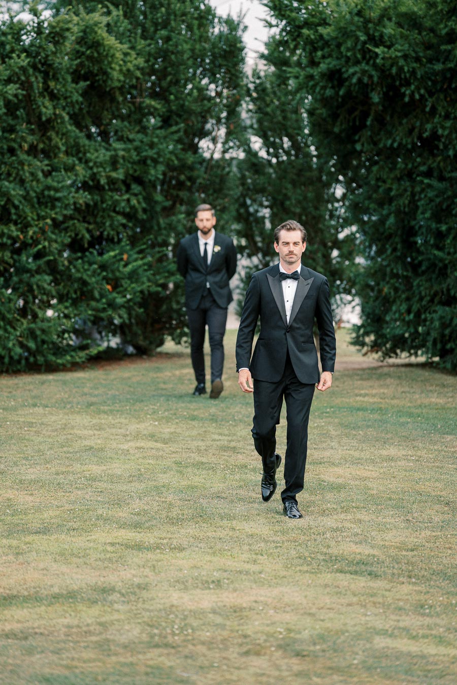 Two men in formal black suits walking on a grassy path lined with tall green trees, capturing a moment of elegance and sophistication outdoors.
