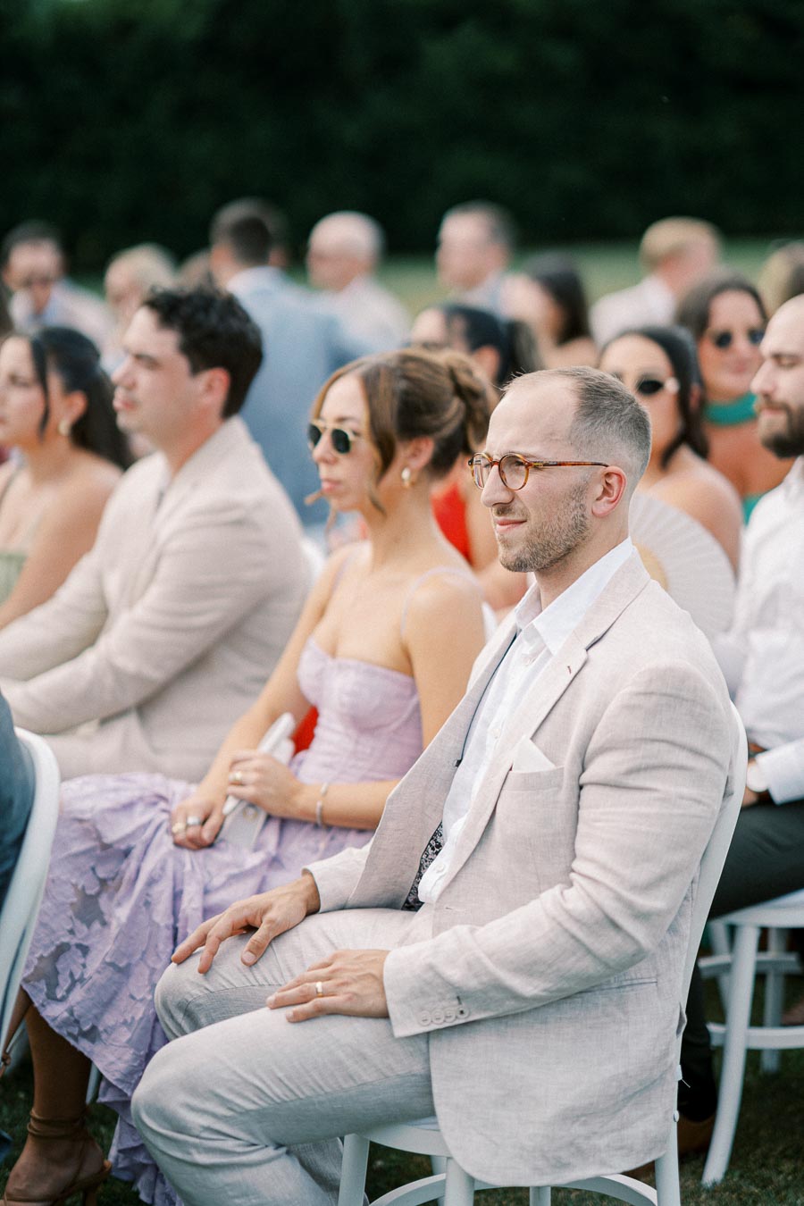 Guests seated outdoors at a formal event, wearing stylish summer attire including a light suit and a lavender dress, attentively watching the proceedings.