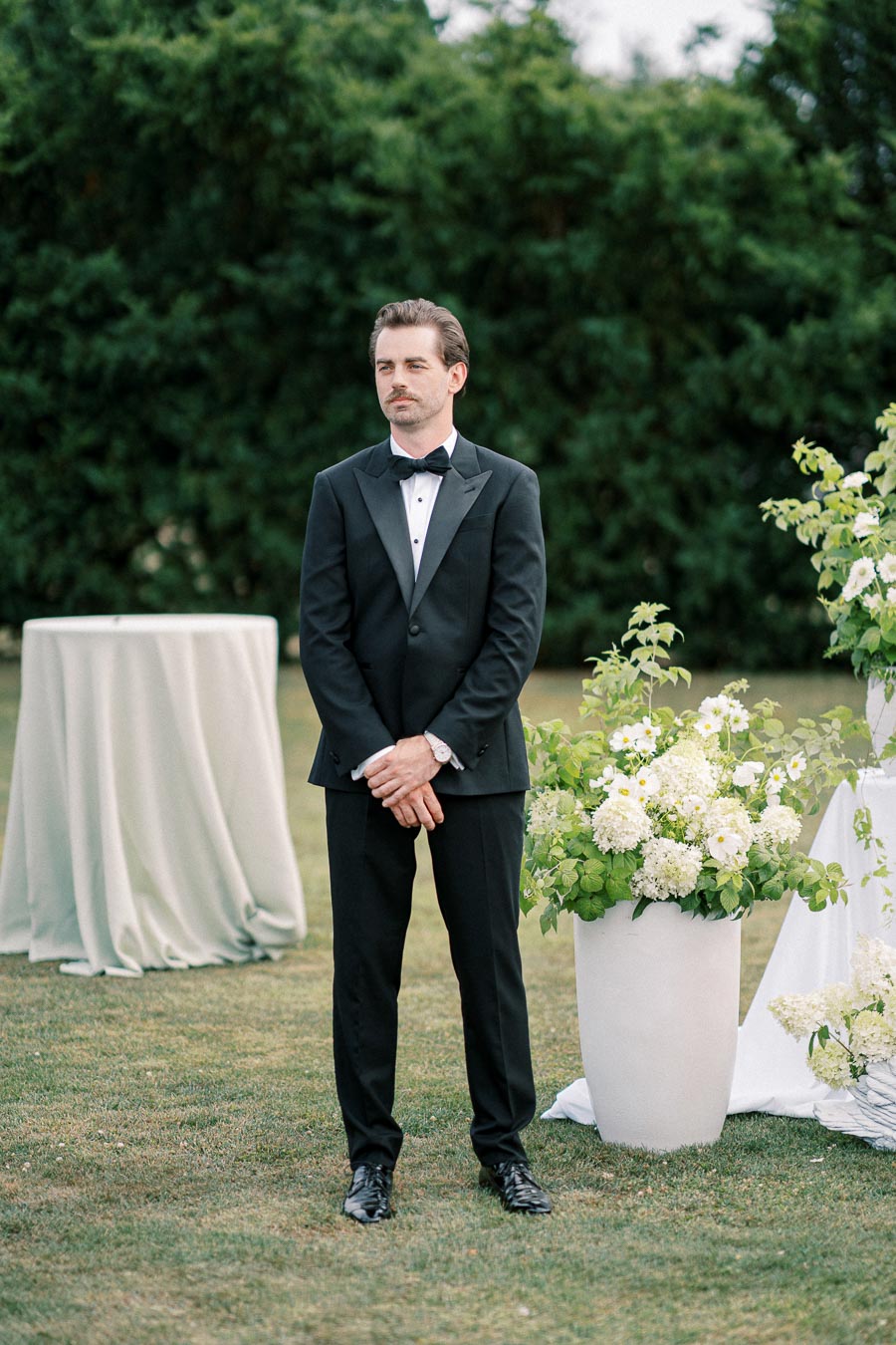 A man in a classic black tuxedo stands on a landscaped lawn, surrounded by lush greenery and elegant arrangements of white flowers, conveying a formal and serene outdoor event setting.