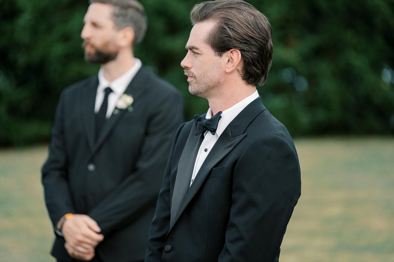 Two men in formal black suits, standing outdoors, captured in profile view against a blurred green background, with focus on the man in the foreground wearing a black bow tie. This image conveys a formal event setting, possibly a wedding or special occasion.