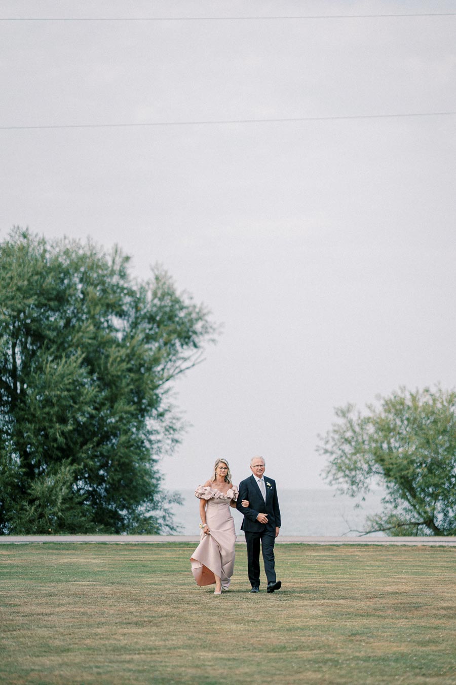 A woman in an elegant pink dress walks arm-in-arm with a man in a suit across a grassy field with green trees in the background.