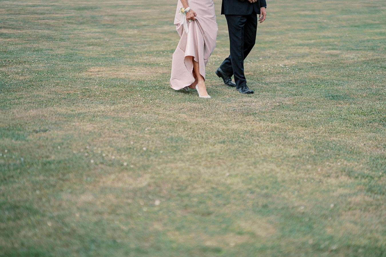 Elegant couple walking on grass, woman in a blush gown holding flowers and a man in a black suit, showcasing wedding or formal attire.