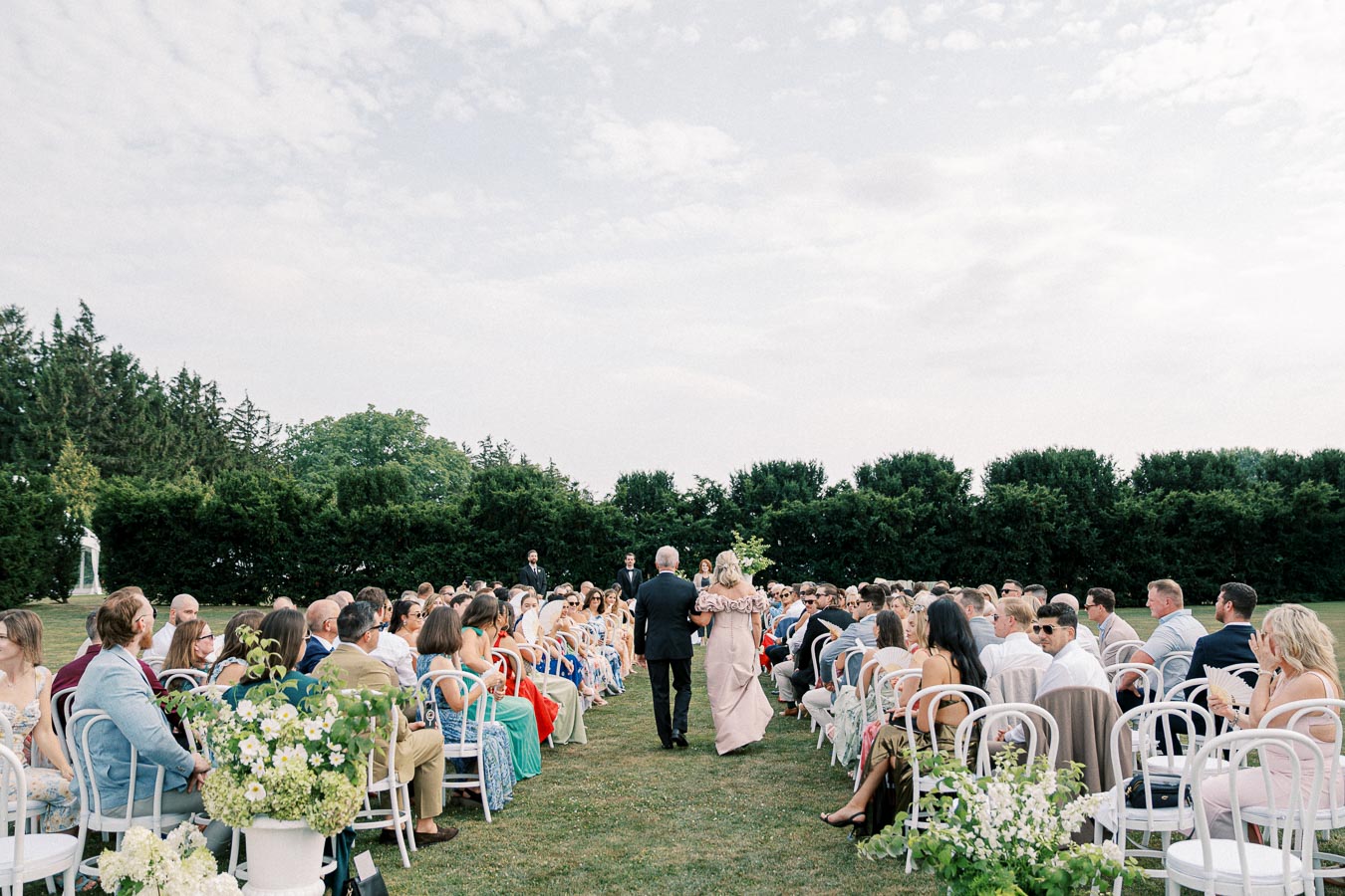 Outdoor wedding ceremony with guests seated on white chairs, lush greenery backdrop, and a couple walking down the aisle.