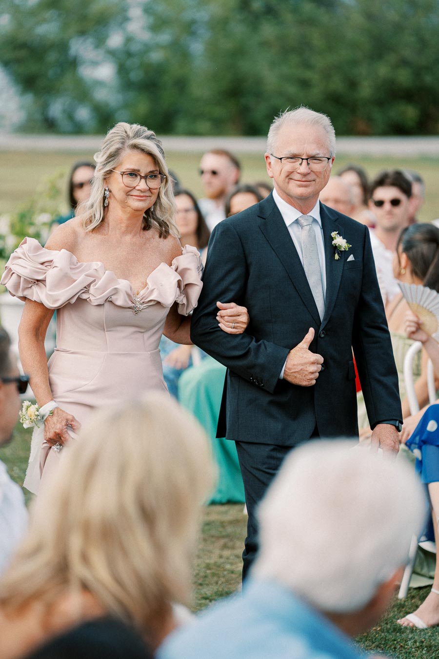 An elegantly dressed couple walks down the aisle at an outdoor wedding ceremony, surrounded by seated guests and greenery. The woman is wearing a pink off-shoulder dress with floral details, while the man is in a dark suit with a light gray tie and boutonniere.