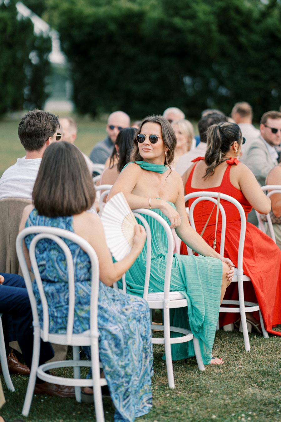 Outdoor wedding ceremony with guests seated on white chairs, featuring a woman in a green dress and sunglasses looking to the side, surrounded by other attendees in vibrant attire on a sunny day.