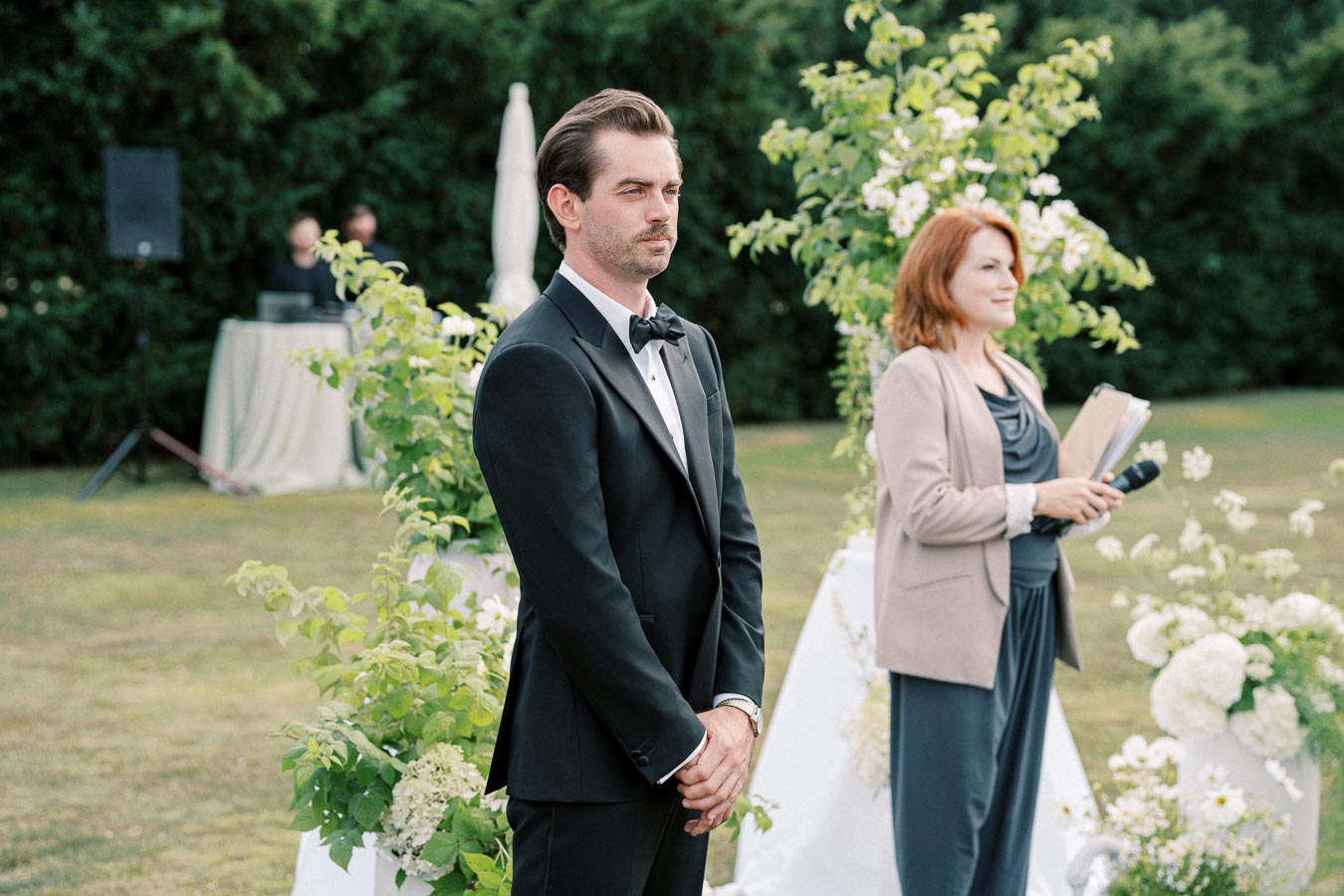 A groom in a black tuxedo stands outdoors next to a woman holding a microphone and notes during a garden wedding ceremony, with green foliage and white flowers in the background.