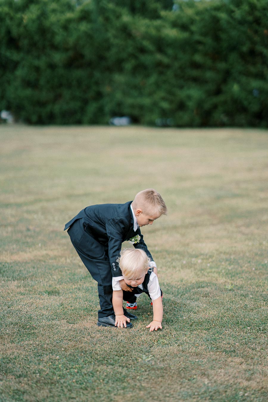 Two young boys in formal attire playing on a grassy field, with a blurred green hedge in the background. The older boy is helping the younger one balance.