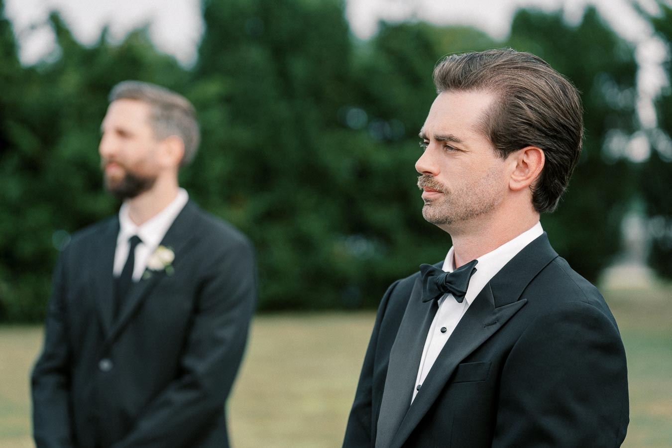 Two men in formal attire, with tuxedos and bow ties, standing outdoors with blurred greenery in the background.