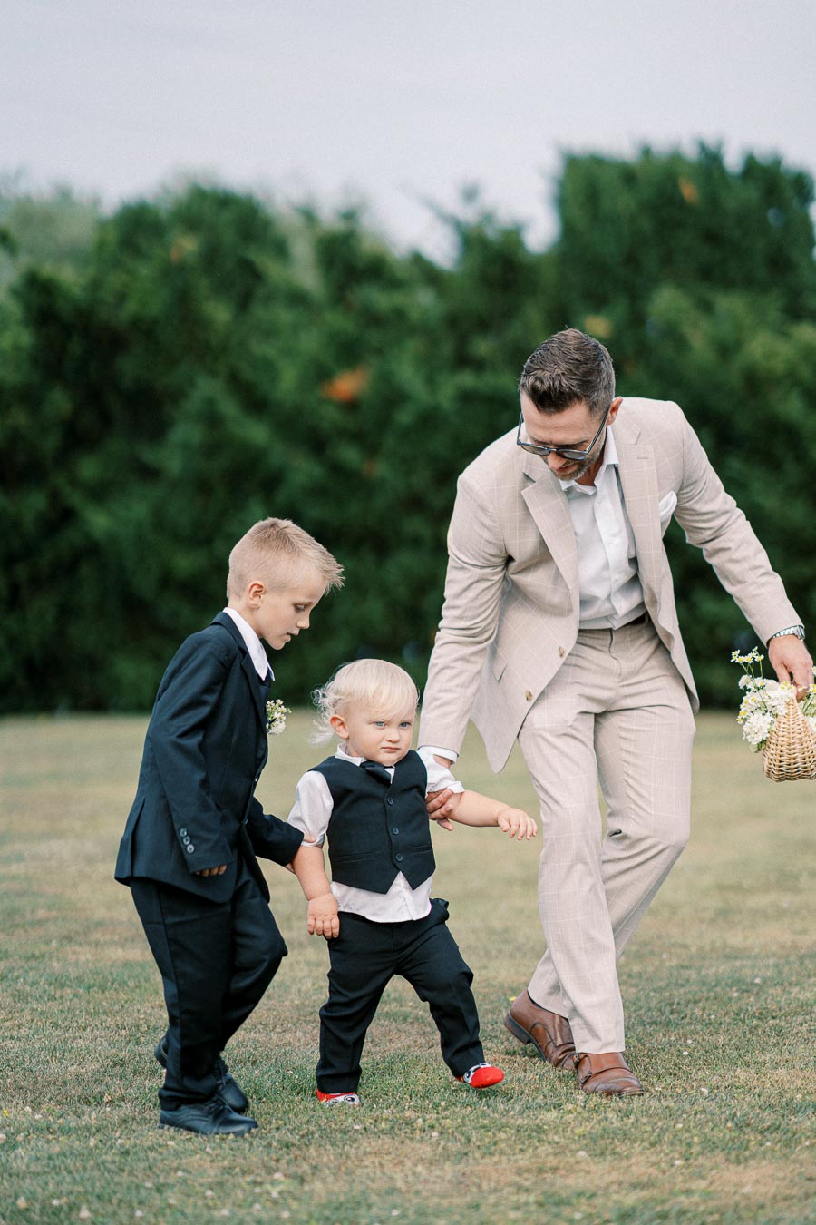 A man in a beige suit helps two young boys in formal attire walk across a grassy area during a wedding ceremony, holding a basket with flowers.
