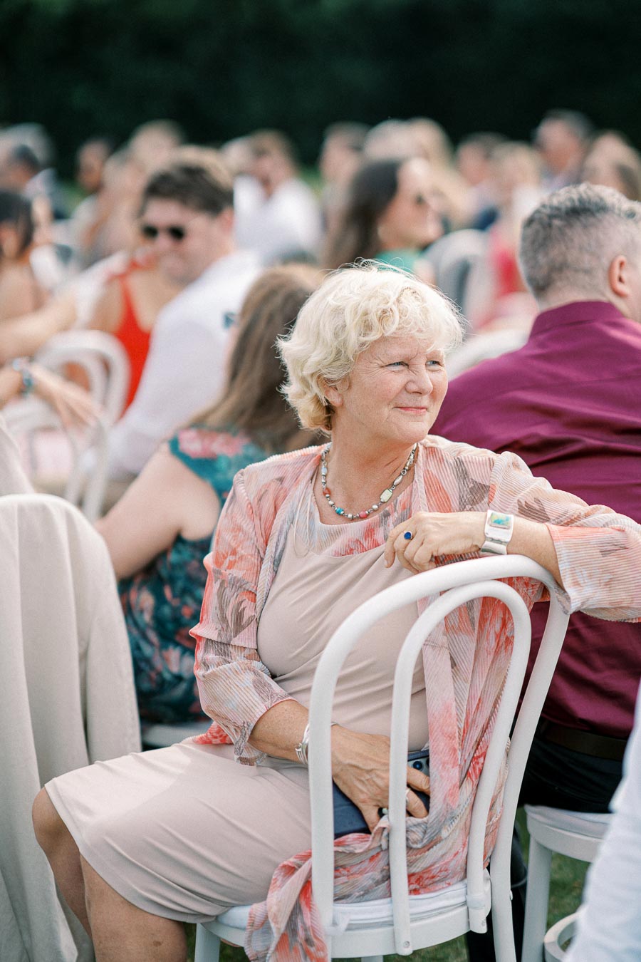 Elderly woman enjoying an outdoor event, sitting on a white chair, wearing a colorful shawl, with attendees in the background.