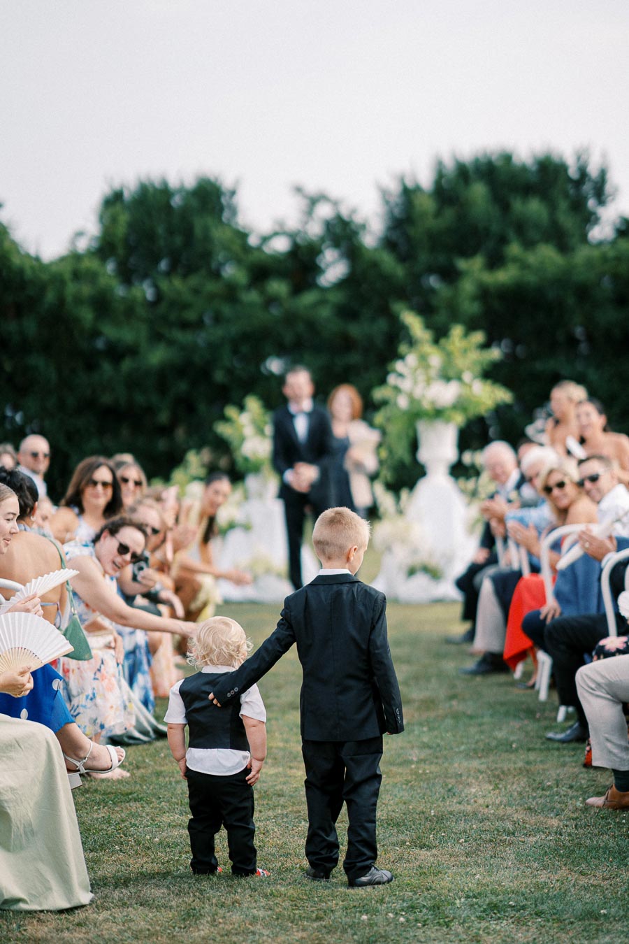 Two young boys in formal attire walk hand in hand down an outdoor wedding aisle, surrounded by seated guests, under a clear sky and lush greenery.