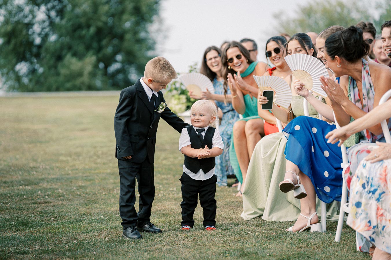Two young boys in suits, smiling and holding hands at an outdoor wedding ceremony, with guests applauding and enjoying the moment.