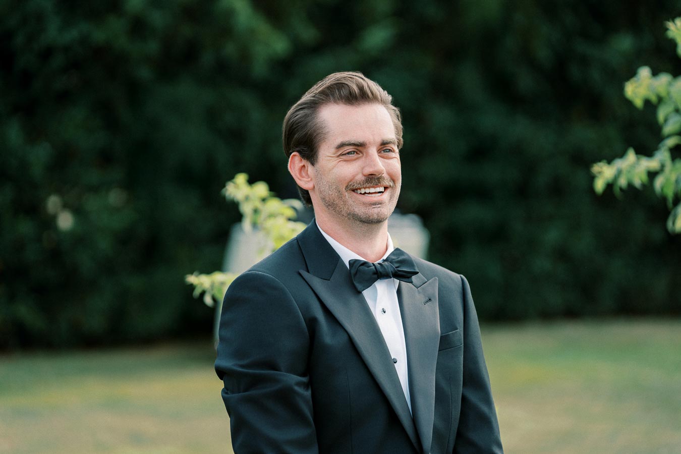 A smiling man in a black tuxedo and bow tie standing outdoors against a blurred green background.
