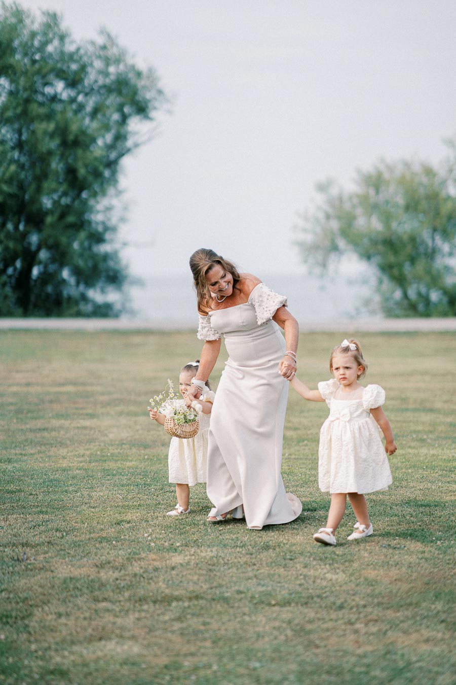 A woman in an elegant off-shoulder dress walking hand-in-hand with two young girls in matching white dresses on a grassy field, carrying a basket of flowers, with trees in the background.