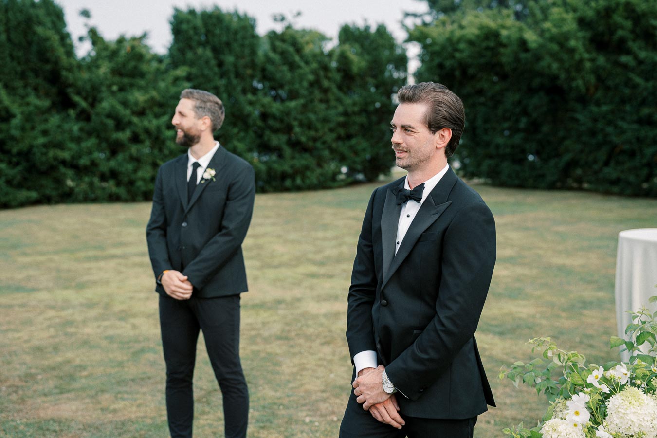 Two men in formal black suits standing outdoors on a grassy area, one looking ahead and the other smiling to the side, surrounded by green foliage and a table with flowers.